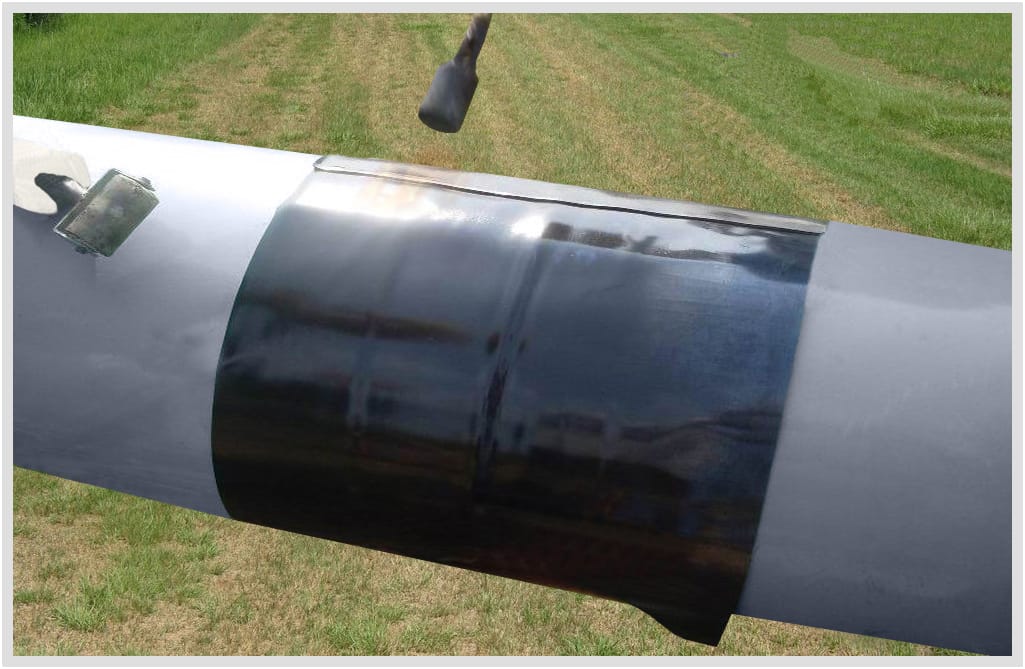 A black three-layer sleeve shrunk over a girth weld on a large grey-coated pipe lying in grass. Its closure patch shows as a raised strip along the top.